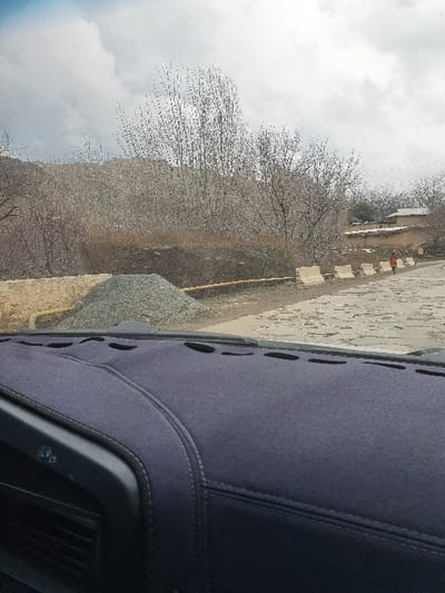The image presents a view from the interior of a vehicle, looking out onto a rural scene in Asmansay, Uzbekistan, under an overcast sky. The foreground reveals a dark blue, textile dashboard cover with stitched seams. Beyond the vehicle's windshield, a paved road, possibly cobblestone or distressed asphalt, curves gently to the right. On the left side of the road, a pile of grey gravel rests adjacent to a low, light-colored stone wall. A series of five light-colored concrete barriers line the right side of the road. A single individual, potentially a child, wearing a reddish-orange garment, walks on the right side of the road, alongside these barriers. The midground features numerous bare deciduous trees and shrubs, indicating a colder season. In the background, several low-rise structures with light-colored walls and flat roofs are visible, situated at the base of hills or mountains also sparsely covered with bare vegetation. The upper portion of the scene is dominated by a cloudy sky.