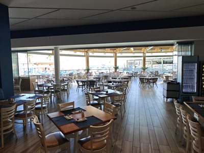 This is a bright, daytime interior shot of a spacious and modern restaurant or dining area, likely part of a resort or hotel in Gouves Municipal Unit, Greece. The room is currently empty of people but is set up for service, with numerous tables and chairs arranged throughout.

The foreground features rectangular wooden tables, some with dark blue or black placemats and cutlery already laid out. The chairs are a mix of white-painted wood and some with wicker backs and seats. The flooring is light-colored wood laminate.

Large glass windows span the far side of the room, offering a panoramic view of an outdoor area. Beyond the windows, sun loungers with blue mattresses and white umbrellas are visible, suggesting a pool or beach area. Palm trees can be seen through the glass, adding to the resort ambiance. The outdoor scene is bathed in bright sunlight, indicating clear weather.

In the mid-ground, a variety of tables and chairs, including round and square configurations, are visible, many featuring wicker designs. Overhead, ceiling lights or fans are integrated into the modern ceiling structure. To the right, a tall, glass-fronted refrigerator is visible, stocked with bottles of water. Faint text on the refrigerator reads "AMINA BEACH." A grey rolling service cart is also present near the refrigerator.

The overall impression is one of readiness and potential activity, a clean and inviting space awaiting its guests, benefiting from ample natural light and a pleasant outdoor vista.