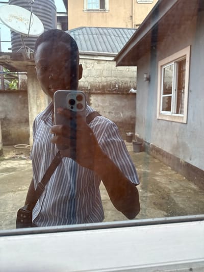 This image captures a person taking a mirror selfie through a window in the city of Iriebe, Nigeria. The subject, a person with short dark hair and dark skin, is positioned centrally, holding a light-colored smartphone with a triple-lens camera system to photograph their reflection. They are wearing a short-sleeved, vertically striped shirt (light and dark grey/blue) and have a dark strap, possibly from a shoulder bag, crossing their chest. The lower portion of a white window frame is visible at the bottom of the image, indicating the reflective surface.

Beyond the reflection, the background reveals an outdoor urban environment. To the subject's right, a grey-rendered building with a white-framed window and interior blinds is visible. A concrete block wall, topped with barbed wire, spans across the midground. Further back, a multi-story building with a light brown facade and a dark corrugated metal roof is present. In the upper left background, a large black cylindrical water tank rests on a concrete support structure, adjacent to a white satellite dish. The ground is a paved or concrete surface, and several dark-colored pots are visible along the base of the grey-rendered building.
