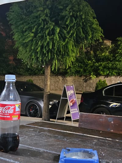 An outdoor scene at night in Biaga, Bulgaria, features a dark-colored table in the foreground. On the table, a nearly empty Coca-Cola bottle with a white cap and a blue ashtray with ash are visible. Behind the table, a large, leafy green tree stands tall, its branches extending upwards. A stone wall and dense green foliage form the background. Two cars are parked behind the tree: a dark gray sedan on the left with its wheels and part of its body visible, and a black sedan on the right. An A-frame sign advertising "Milka" chocolate, featuring an image of chocolate ice cream, is positioned between the cars. The lighting suggests artificial illumination, likely from streetlights or nearby buildings, casting shadows and highlighting certain elements of the scene.