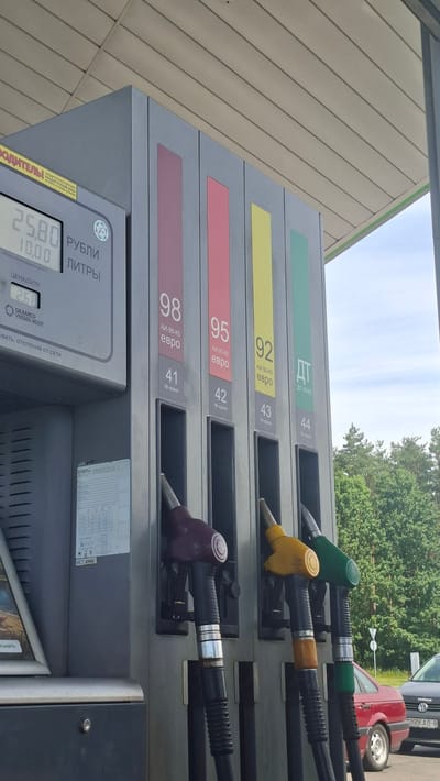 This media file shows a close-up of a gasoline pump at a service station. The pump has four different fuel nozzles, each labeled with the octane rating and type of fuel: 98, 95, 92, and DT-L-K5. The prices are displayed on the pump's digital screen, showing 25.80 rubles for volume and 2.68 for price per liter. The context indicates the location is in the Drachkovskiy rural council, Belarus.
The scene appears to be outdoors, with natural light suggesting daytime. The sky is visible in the background, partially obscured by trees. There are vehicles in the background, including a red car and another partially visible car. The overall setting is a typical gas station forecourt.
The focus is on the fuel pump and its display, with the nozzles and price information being the primary subjects. There are no people clearly visible in the frame, but the presence of the gas pump and vehicles implies a transaction or refueling activity is taking place or has recently occurred. The weather appears to be fair. The visible text includes fuel types, octane ratings, prices, and manufacturer information on the pump.