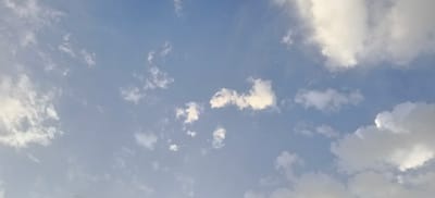 This is an aerial photograph of a partly cloudy sky, taken in Margilan City, Uzbekistan. The sky is a pale blue, with scattered white clouds of varying sizes and densities. Some clouds are wispy and thin, while others are more dense and fluffy. The sun appears to be behind some of the clouds, creating areas of brightness and shadow. There are no people, structures, or any other objects visible in the image, only the sky and clouds.