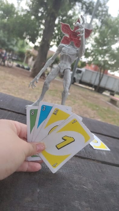 A person holds Uno cards with the numbers 1, 6, 3, and 8 displayed in yellow, blue, and green. In the background is a Demogorgon figure, a truck, trees, and a picnic table. The image is slightly blurred with an off-center angle.