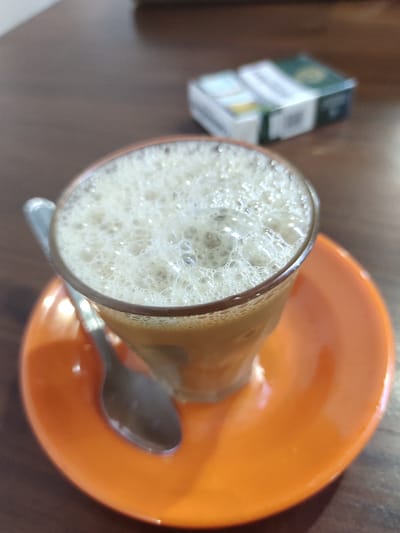 A close-up, eye-level shot shows a foamy beverage in a clear glass on an orange saucer. A spoon rests on the saucer next to the glass. In the blurred background, a pack of cigarettes sits on a wooden surface. The setting appears to be a cafe or restaurant in Kutalhoksukon, Indonesia.