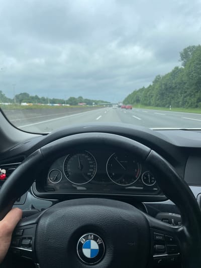 The image displays the interior perspective of a BMW automobile from the driver's seat, situated on a multi-lane highway in Münster, Germany. A human hand is visible on the left side of the black leather-wrapped steering wheel, which features the prominent BMW logo at its center and integrated controls for cruise control, media, and telephone functions. The instrument cluster is visible through the steering wheel, showing a speedometer with the needle indicating approximately 120 km/h, a tachometer, and gauges for fuel level and engine temperature. A digital display within the cluster indicates an ambient temperature of "17.0 °C", time "12:50", "D" for drive mode, "014 Km", and "3971.1" (likely odometer reading). A small, red, ladybug-shaped decoration is attached to an air vent on the left dashboard.
The vehicle is moving forward on a wet, asphalt highway, suggesting recent rainfall. White dashed lines delineate the road lanes. In the distance, several vehicles, including red passenger cars and at least one truck, are visible on the same carriageway. Both sides of the highway are bordered by dense green trees, forming a wooded landscape. The sky is uniformly overcast with light grey clouds, indicating a lack of direct sunlight. The overall environment is consistent with a German Autobahn near the city of Münster.