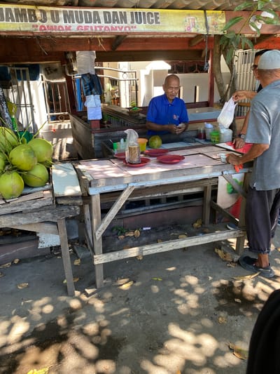 A street vendor in Santan, Indonesia, is serving a customer at an outdoor stall. The vendor, a bald man wearing a blue polo shirt, stands behind a wooden counter laden with fresh coconuts, plates, and jars. He is in the process of accepting payment from a customer, a man in a gray shirt and a white skullcap, who is handing him a white plastic bag. Another person, mostly obscured, stands behind the customer. The stall is adorned with a sign that reads "JAMBU U MUDA DAN JUICE" and "AWAK GEUTANYO". The scene appears to be in an open area with dappled sunlight filtering through trees, creating shadows on the concrete ground. The overall impression is one of a casual, local transaction.