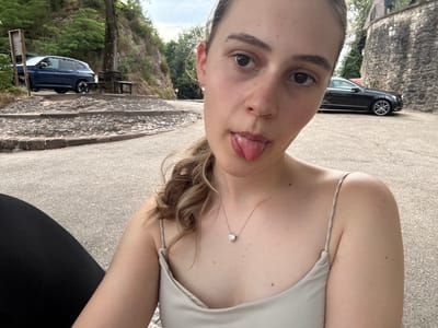 A young woman with brown hair pulled back in a ponytail, and wearing a light-colored slip dress and a heart-shaped pendant necklace, sticks her tongue out at the camera. She is seated outdoors on a paved surface with a blurred background of a rocky hillside with trees and cars. A dark blue SUV is parked on an elevated paved area to the left of the frame. A black Mercedes-Benz is parked on the right, next to a stone wall. The scene appears to be a scenic overlook or roadside rest stop.