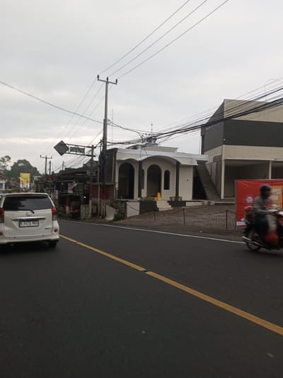 The image shows a street scene. A white car with the license plate B 1473 NKX is on the left side of the road. On the right, a person is riding a motorcycle. A building with arches and a dome sits next to a two-story structure, with a banner partially visible on the second building. Power lines stretch across the sky. The road has a yellow line marking. Small shops or stalls are also visible in the background. There is no information in the image that this is Taunggyi, Myanmar.