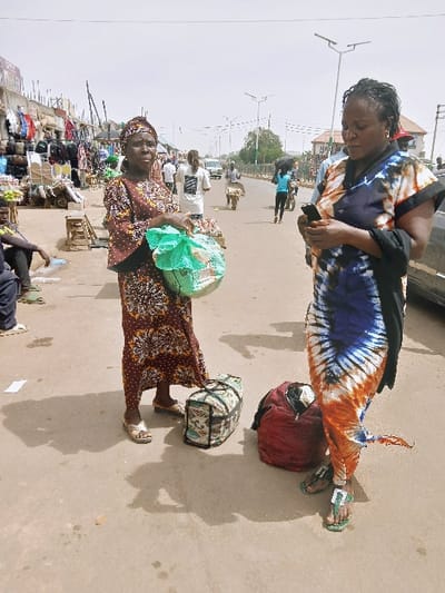 The image depicts an outdoor street scene in Kwekwe, Zimbabwe. Two adult females are prominent in the foreground. The woman on the left, dressed in a patterned brown and cream long dress with a matching headwrap and light sandals, holds a green plastic bag in her left hand and looks towards the viewer. A patterned duffel bag rests on the paved ground near her feet. To her right, a second adult female, wearing a blue, white, and orange tie-dye patterned dress and green flip-flops, holds a black smartphone horizontally with both hands, looking down at the device. A red bag rests on the ground near her feet.
The background shows a busy urban street extending into the distance under a clear sky. Stalls displaying various merchandise, including clothing and bags, line the left side of the street. Numerous other individuals are visible further down the street, some walking, others operating pushcarts or near vehicles. A white vehicle is partially visible on the street, along with several streetlights featuring rectangular solar panels. The ground is a light-colored, possibly dusty, paved surface with long shadows cast by the people, indicating strong daylight.