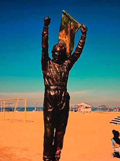 A full-body bronze statue of a person holding a flag aloft stands on a sandy beach. The statue is dark brown and appears to be made of weathered bronze. The person is wearing what looks like a uniform or athletic attire, with long sleeves and pants. Their right arm is raised, holding a flag that is partially unfurled and appears to be made of a metallic material, possibly brass, with patches of rust and discoloration. The flag's design is not clearly discernible.
The statue is positioned on a wide, sandy beach, with the ocean visible in the background. The water is a light blue, meeting a pale horizon. The sky above is a vibrant, clear blue.
In the mid-ground, a yellow soccer goal is visible on the left side of the frame. Further back, a white canopy structure with "SCUL RIO 15" written on it is present, with people gathered around it. Several beach umbrellas, some colorful and some striped, are scattered across the sand. A few people are seated on chairs under these umbrellas, and others are walking or standing. The overall atmosphere suggests a sunny day at a popular beach.
The location is identified as the city of Natal, Brazil, specifically on Ponta Negra beach, a well-known tourist destination famous for its sand dunes and vibrant atmosphere. The statue itself is a landmark, often associated with the spirit of victory and sport, given its location and the celebratory pose.