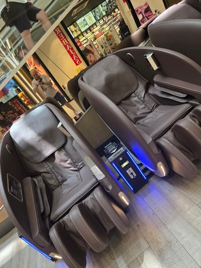 The image displays two brown leather massage chairs, with one in the foreground and another behind it. Both chairs have the "RELAXA" logo visible on their headrests. In the background, there is a retail environment with shelves stocked with various products, including cosmetics and beauty items. A red banner with white text reading "REBALA" is also present. To the left of the chairs, a man is visible in the reflection of a glass surface, holding a smartphone and taking a picture. He is wearing a white t-shirt and dark shorts. The floor is made of light-colored wood planks. Blue LED lights are illuminating the base of a control panel situated between the two massage chairs, which appears to be a payment or timer unit. The overall setting suggests a commercial establishment, possibly a mall or a dedicated store for massage chairs. The specific location in Collado Villalba, Spain, is not identifiable from the visual cues within the image itself.
