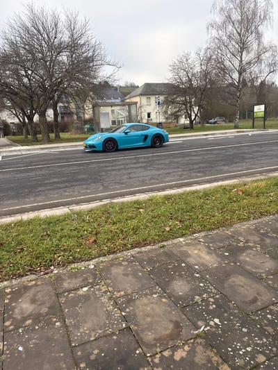 This image captures an outdoor scene in Bech-Kleinmacher, Luxembourg, under an overcast sky. A light blue (turquoise) Porsche Cayman two-door sports car is prominently featured in the mid-ground, positioned on an asphalt road and oriented towards the right of the frame. The vehicle has black multi-spoke wheels and red brake calipers, with a yellow European license plate visible on its front bumper. The road, dark gray, has white solid lines marking the edges and a dashed white center line.
In the foreground, a sidewalk made of rectangular, dark gray concrete slabs shows signs of lichen growth and small plants emerging from cracks. Between the sidewalk and the road lies a narrow strip of sparse green grass with scattered brown leaves.
The background includes numerous leafless deciduous trees, suggesting a winter or early spring season. Behind the road, several buildings are visible, including a prominent light-colored, multi-story structure. A glass-paneled bus stop shelter is situated to the left of this building, on the far side of the road. A blue and white sign is attached to a pole near the main building, and further to the right, a green informational sign on a stand is present in a grassy area. A dark-colored SUV is partially visible in the far background, positioned on a distant road or lot. No individuals are visible within the frame.