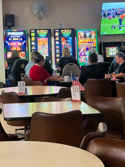 The image depicts the interior of a VFW hall in Atlantic Beach, Florida, specifically the bar area, as evidenced by the presence of slot machines and a sign that reads "POST 316." The hall is set up for a gathering, likely related to the VFW, with slot machines lining the back wall. Several people, predominantly older adults, are seated and engaged with the machines, with some interacting socially. The lighting is dim, with the bright, colorful displays of the slot machines creating a vibrant atmosphere. The foreground shows tables with chairs, set up for dining or socializing, with American flags and table numbers visible, suggesting a special event or tournament. The overall impression is a casual, community-oriented space where patrons can enjoy entertainment and social interaction.