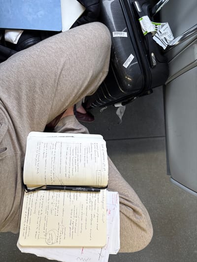 The image is a close-up, high-angle shot of a person's lap in what appears to be a public transport setting, possibly an airport or train station, as indicated by the luggage. The person is wearing light brown sweatpants. On their lap rests an open notebook with handwritten notes, and a black fine-tip pen is positioned across it. To the right of the notebook is a black suitcase with luggage tags attached. In the background, to the left, a portion of a light blue laptop is visible, along with black clothing. The overall scene suggests a moment of study or work in transit. There is no information in the image that places the scene in Reigate and Banstead, United Kingdom.