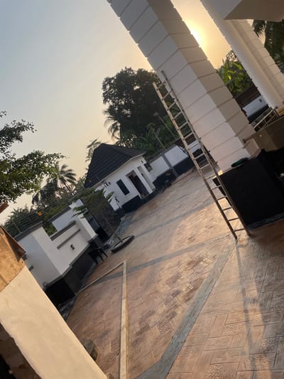 The scene depicts an outdoor paved courtyard within the city of Ogbunu, Nigeria, illuminated by low-angle sunlight, indicative of either sunrise or sunset. A large, multi-segmented white pillar stands prominently on the right, with a silver-colored extension ladder propped against its lower section. The ground consists of patterned, light brown stamped concrete, featuring alternating wood-grain and geometric textures, segmented by dark linear borders, with strong sunlight reflecting off its surface.
In the midground, a small, white single-story building with a dark, pyramid-style roof and dark rectangular window or door openings is visible. Perimeter walls, white with dark bases and decorative horizontal bands, enclose the area. Tropical trees, including several palm trees, are densely clustered in the background and along the fence line. A small sapling planted within a black tire planter is situated on the paved area. A dark stool or low table is positioned near the base of the white wall. A large, dark, rectangular outdoor unit rests near the base of the prominent white pillar. A long, light-colored frame lies diagonally across the paved surface in the lower-center. No individuals are present, nor are any active interactions or specific actions depicted within the frame.