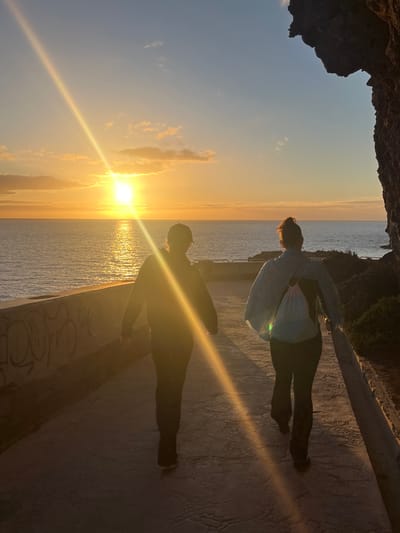 A person in a light-colored shirt and dark pants stands on a textured concrete surface with graffiti along the edge. The sun is setting over the ocean, creating a golden glare and a bright reflection on the water. The silhouette of another person is visible on the concrete surface, along with the person's shadow. The sky is a gradient of blues and yellows. A rocky overhang appears in the upper-left corner of the image. Lush greenery is visible along the upper-right edge. The image appears to have been taken from a high vantage point overlooking the ocean, at sunset. Puerto Rico is located in Gran Canaria, Spain.