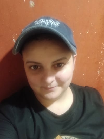This is a vertically oriented, close-up, over-the-shoulder selfie of a person wearing a black t-shirt and a dark blue baseball cap. The person's face is the primary subject, with their right eye and part of their left eye visible. They have dark hair and a neutral expression. The baseball cap has white embroidered text or a pattern on the front, though it is largely obscured by the angle. The background is a solid, reddish-orange surface, possibly a wall, with some scattered white specks, which could be paint chips or dust. The lighting appears to be artificial and low, casting shadows and highlighting certain features of the face and clothing. The image quality is moderate, with some graininess. There is no indication of the time of day, weather, or specific location beyond the provided context of Chutove, Ukraine. The person is not actively engaged in any discernible activity.