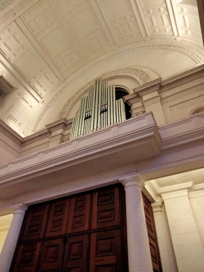 A low-angle shot shows the interior of a grand building. High above is a pipe organ, its pale pipes against an arched window space. Ornate moldings surround the organ. Below, a large, dark wooden door fills the frame. Two white columns, one on each side, flank the door. The ceiling features a textured, patterned design. The overall color palette is dominated by light beige and dark brown.
