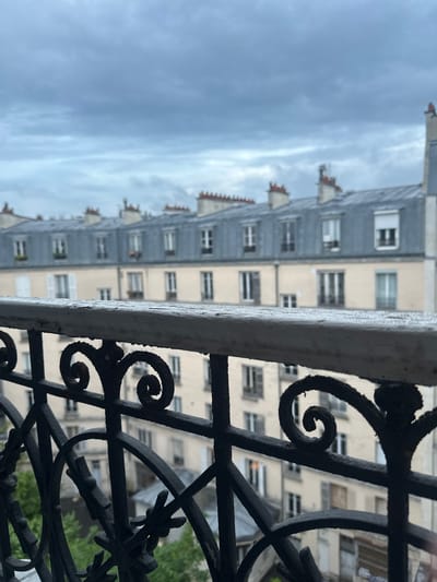 A medium shot captures a view from a balcony in Saint-Ouen-sur-Seine, France. The foreground is dominated by an ornate black wrought iron railing, its design featuring prominent swirls and decorative elements. A white, rectangular support beam runs horizontally across the frame, slightly obscuring the view beyond.
In the background, a Parisian building is visible, characterized by its light beige facade and rows of windows. Above the building, the sky is overcast with gray clouds. The overall perspective is slightly tilted to the left.