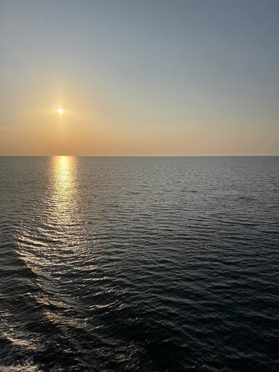 The image captures an expansive view of open water under a clear sky, positioned in Kato Karioutes, Greece. The horizon line is sharply defined, dividing the upper sky from the lower sea. In the upper left quadrant, a bright, circular, yellow-white sun emits a prominent glow, extending as a shimmering, elongated golden reflection across the water towards the lower left of the frame. The sky transitions from a light blue at the top to a warm orange-yellow gradient closer to the horizon. The sea surface is characterized by small, uniform ripples, with the area directly impacted by the sun's reflection appearing brighter and more defined, while the remainder of the water exhibits a darker blue-grey tone. No land, vessels, individuals, or other specific objects are present within the visible frame.