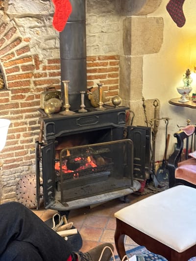 This media file captures an indoor scene, likely in a cozy room or restaurant in Étréchy, France, featuring a traditional fireplace. The central focus is a black, cast-iron wood-burning fireplace or stove, with a lively fire glowing brightly behind a protective mesh screen. A dark, metallic flue pipe extends upwards from the stove, against a rustic wall composed of rough-hewn stone and red brickwork.

Various decorative and functional objects adorn the space. To the left of the fireplace, several brass or gold-toned ornamental pieces are arranged. A set of fireplace tools, including a poker and tongs, leans against the wall to the right of the stove. Above, a section of the wall is painted white, with a small shelf holding a decorative bowl or vase. Festive touches include a red and black plaid stocking hanging from the upper left, and another red textured decoration further down the wall.

On the right side of the image, part of a wooden chair with a white upholstered seat is visible. Below it, the lower legs and feet of a person are seen, clad in dark trousers and shoes with red-patterned socks, suggesting they are seated and enjoying the warmth. Small pieces of kindling are scattered on the terracotta-tiled floor near their feet, and a circular, perforated metal grate sits in front of the fireplace.

The scene evokes a sense of warmth, comfort, and relaxation, with the glowing fire creating a welcoming ambiance. No visible text is present in the image.