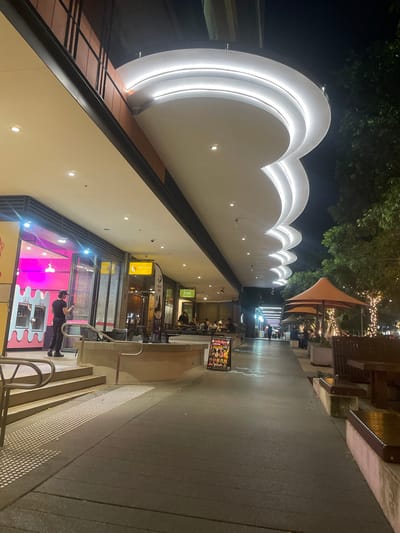 The image depicts a nighttime view of a pedestrian walkway in Brisbane, Australia. The walkway is lined with shops on the left and outdoor seating areas on the right. A distinctive architectural feature is a wave-like canopy with integrated lighting extending over the walkway. On the left, a person in a black uniform is standing in front of a brightly lit shop with pink interior lighting. To the right, there are several outdoor dining tables and chairs, some under orange umbrellas, with decorative string lights illuminating the trees and surrounding area. People are seated at some of the tables. The walkway is paved and has tactile paving indicators near the steps leading up to the shops. A sign with colorful images and text stands near the walkway. The overall impression is of a lively, well-lit urban entertainment precinct at night.