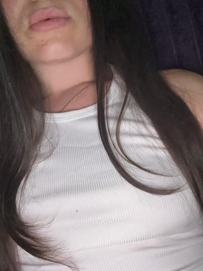 The image is a close-up shot of a person's upper body, focusing on their face and chest. The individual has long, dark hair that frames their face and falls over their shoulders. They are wearing a white, ribbed tank top. A thin silver necklace is visible around their neck. The background is dark and appears to be indoors. The person's lips are slightly parted, and their chin is visible. There is a small mole on their chest. The image does not provide any information about the location in the city of Bucha, Ukraine, or any specific actions or interactions.