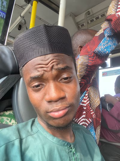An image taken inside a public transport vehicle, likely a bus, in Ebute-Metta, Nigeria, captures a close-up selfie of a young Black male with other passengers in the background. The primary subject, appearing to be in his late teens or early twenties, occupies the center-left foreground. He wears a dark green collared shirt with visible buttons and a black, textured kufi cap featuring a geometric pattern. His expression is neutral to slightly contemplative, with a furrowed brow, and he has a faint beard and mustache.
Behind and to the immediate right of the primary subject, a second male passenger is partially visible from the back, showing the top of his head with short, dark hair. He is wearing a long-sleeved garment with a vibrant, multi-colored geometric pattern in red, blue, yellow, and white, consistent with Ankara fabric. Further to the right and slightly behind, a third passenger is partially visible, wearing a dark maroon or purple garment, with an arm holding an indistinguishable dark object.
The interior environment features a white ceiling, a yellow vertical grab pole, and a white vertical support pole. Grey seat backs are partially visible on the left. Through a window on the right, a white sign with black text, partially obscured, is visible, possibly including phrases such as "CASH payments" and "police." On the far left, an external view through another window shows hints of green foliage and a sign displaying the word "CITY." The overall scene suggests a routine commute within an urban setting.