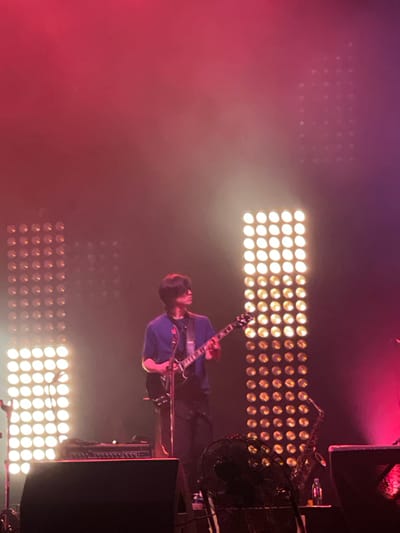A musician is playing an electric guitar on a stage in Salembaran, Indonesia. Behind the musician are several panels of lights and a fan. A large speaker is visible to the right. The scene is lit with bright white stage lights and red lights.