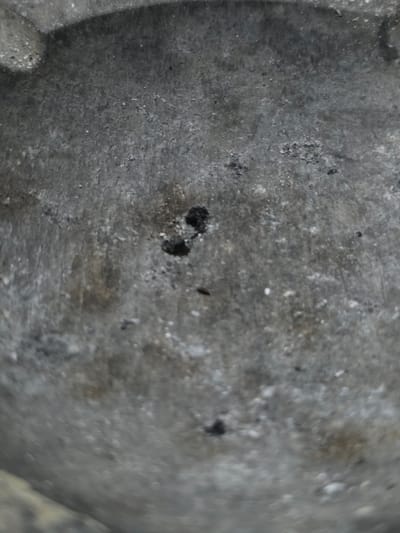 The image shows a close-up, slightly out-of-focus view of a textured, gray, circular object. The surface appears to be metallic or stone, with a rough, granular texture and some lighter flecks. There are several small, dark specks scattered across the surface, and two particularly noticeable dark specks close together in the upper central portion of the image. The lighting suggests a diffused, overhead light source. The image does not contain any information that would allow for its placement within the city of Bireuenmeunasahblang, Indonesia.