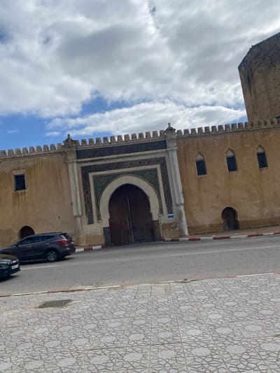 The image displays a segment of a fortified wall featuring a prominent gate, located in Mechouar Fes Jdid, Morocco. The wall, constructed of light brown material, is topped with battlements. To the left of the main gate, a single rectangular window is positioned high on the wall, with a smaller arched doorway at ground level partially obscured by a vehicle. To the right of the gate, three smaller arched windows are visible, along with another arched doorway at ground level. A large, light brown defensive tower partially extends into the frame from the far right.
The central feature is a large, dark brown, arched wooden double door, set within an elaborately decorated archway. This archway is framed by white architectural elements and adorned with intricate geometric patterns in dark blue/green, white, and brown.
An asphalt road occupies the mid-ground, separated from the wall by a red and white painted curb. A dark-colored SUV is parked on the street to the left, partially obscuring the lower left portion of the wall. The front of a second dark vehicle is visible at the extreme lower left.
The immediate foreground consists of a grey tiled sidewalk featuring a repeating star-patterned geometric motif. An irregular dark stain is present on the sidewalk. The sky above is partly cloudy, displaying white and grey clouds against a blue background. No individuals, specific actions, or interactions are depicted within the scene.