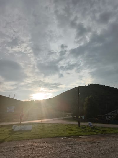 The image depicts a landscape scene in the city of Aya, Russia, likely during the early morning or late evening given the low sun angle and dramatic lighting. The foreground consists of a gravel path with patches of green grass and some concrete barriers. In the middle ground, a road curves around the base of a hill, which is covered in trees and vegetation. The sun is low on the horizon, creating a strong lens flare that illuminates the sky with bright white and yellow light. The sky itself is filled with a mix of gray and white clouds, some of which are illuminated by the sun. A few utility poles and wires are visible, along with a sign on the left side of the road. In the distance, a few houses with dark roofs are nestled among the trees on the right side of the frame. The overall atmosphere is serene and slightly hazy due to the sunlight.