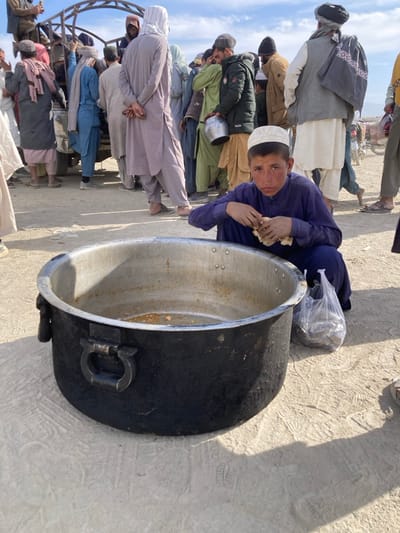 This image, set in an unpaved outdoor area of Chaman City Tehsil, Pakistan, depicts a scene likely involving food distribution or a communal gathering. In the foreground, a young boy, approximately 8-12 years old, wearing a purple shalwar kameez and a white kufi cap, squats beside a large, empty cooking pot. He is actively eating a piece of light-colored flatbread. To his immediate right, a clear plastic bag filled with dark, granular items rests on the dusty ground. The large pot, black on its exterior with a metallic silver interior, shows residue on its inner surface, suggesting recent use. The background features a crowd of approximately 15-20 adult males, dressed in traditional Pakistani attire including shalwar kameez, turbans, and head coverings. They are standing closely together, some appearing to be in a queue or gathered around what resembles the rear of a white utility truck with a metal canopy frame. One man in the middle ground holds a small silver pot. The environment is characterized by dry, light-colored earth with visible footprints and tire tracks. The sky is clear blue with sparse white clouds, indicating strong daylight and casting distinct shadows.