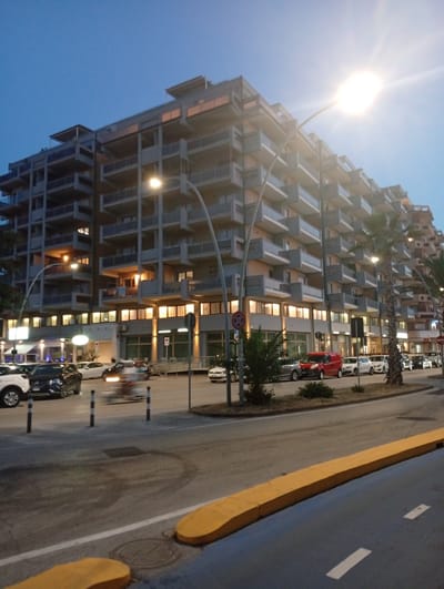 The image shows a wide, multi-story modern building with balconies, illuminated windows, and a flat roofline, situated on a street in Civitanova Marche, Italy. The building is tall and appears to be residential or mixed-use, with a contemporary architectural style featuring horizontal lines and grey facades. Several palm trees are visible in front of and beside the building. The street in front of the building is paved and appears to be a road with parking spaces and a sidewalk. Several cars are parked along the street, and some are in motion, indicated by motion blur. A street lamp with a bright light is prominently featured in the sky, suggesting it is either dusk or dawn, or a very bright street light. The sky is a clear, pale blue, transitioning to a darker shade at the top. The overall scene depicts an urban environment with a focus on the architecture and street life.