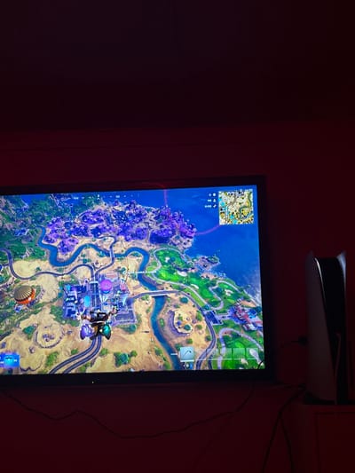 The image captures a close-up view of a television screen displaying a video game, set against a dark red wall. Above the television, a white PlayStation 5 console is mounted horizontally.

The television screen shows a colorful, detailed map from the video game Fortnite. The map depicts a diverse landscape including a large body of blue water on the left, a purple-hued island or landmass, green forested or grassy areas crisscrossed by rivers and roads, and some lighter, possibly desert-like terrain towards the bottom right. Several structures, such as buildings and what appears to be a golf course, are visible within the green areas. A player character, identifiable by its distinct outfit and a purple glider or umbrella, is seen descending through the air onto the map, currently over a more urbanized section with multiple buildings.

In the top-left corner of the screen, a mini-map provides a zoomed-in perspective of a smaller section of the game world. Below the mini-map, partially visible game UI elements include three circular icons, two of which clearly show the number "0," likely indicating resource counts or similar game statistics. A prominent red line extends diagonally across the main map from the top-left, indicating a path or trajectory, possibly of the game's battle bus or a marked route. Small icons and markers are scattered across the map, suggesting points of interest or objectives.

The PlayStation 5 console is mounted just above the top edge of the television, its distinctive white panels and black core clearly visible. Black cables are seen trailing from behind the console and down the wall on the right side of the screen. The wall itself is a uniform, dark red or maroon color. The overall lighting in the room appears dim, with the screen providing the primary source of illumination, suggesting a nighttime or low-light gaming environment.