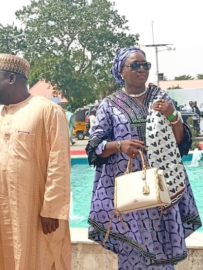 An outdoor scene in Unguwan Sanusi, Nigeria, depicts two individuals in traditional attire by a body of water, likely a fountain or pool. The bright daylight suggests a sunny day.

On the right, a woman stands prominently, facing slightly away from the camera. She wears a striking long-sleeved traditional dress in purple with black patterns and embroidery, complemented by a matching headwrap. Her accessories include large dark sunglasses, a white beaded necklace, white pearl earrings, a gold watch, a gold ring, and white beaded bracelets. A green wristband is visible on her right wrist. She holds a cream-colored handbag with gold hardware and has a white scarf with black, stylized patterns draped over her left arm. Recognizable text on the scarf includes "ZARA" and repetitive letter-like designs, possibly "N" or similar glyphs.

To her left, partially visible, stands a man in a light beige or cream-colored traditional outfit, possibly an agbada, featuring subtle vertical patterns. He wears a patterned, ornate cap. His right hand is visible by his side.

In the background, a light blue fountain or pool adds a vibrant element to the scene. Beyond this, scattered trees and faint outlines of buildings are visible. Further back, vehicles, including a yellow auto-rickshaw (keke napep), and other people can be seen, suggesting a public gathering or event. A tall pole with cameras or antennae is also in the distance. The overall impression is that of a social occasion in a public space.