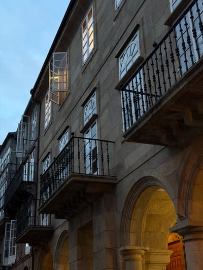The image captures the facade of a historic building in Santiago de Compostela, Spain, likely in the old town, given the architectural style. The building is constructed with large, rough-hewn stone blocks, giving it a weathered and ancient appearance. Multiple windows with white frames are visible, some of which are open, allowing a warm, yellow light to emanate from within, suggesting interior illumination. Several balconies with ornate black metal railings jut out from the facade, adding a touch of elegance to the robust stone structure. The ground floor features a series of arched openings, also framed in stone, with one arch revealing a glimpse of a wooden door and a warmly lit interior. The sky above is a dusky blue, indicating either dawn or dusk. The overall impression is one of timeless architecture bathed in soft, inviting light.