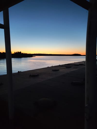 A body of water stretches across the horizon at dusk, reflecting the gradient of the sky from deep blue to orange. Silhouetted trees line the distant shore, creating a dark, jagged edge against the lighter hues of the sunset. In the foreground, a dark, textured surface with several circular, raised objects is visible, suggesting a deck or platform. Two white vertical structures frame the scene on the left and right, adding a sense of depth and perspective. The overall impression is a tranquil, serene evening by the water, possibly from a boat or a pier in Irdomatka, Russia, given the provided context.