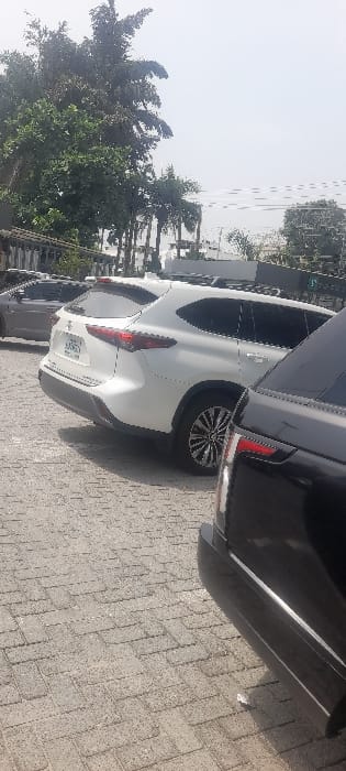 The image displays an outdoor scene in Falomo, Nigeria, characterized by a paved surface and multiple vehicles under bright daylight. The ground is composed of grey interlocking concrete pavers. In the immediate foreground on the right, a partial view of a dark-colored SUV's rear bumper and taillight is visible. Adjacent to it, centrally positioned, is a white Toyota Highlander SUV, observed from its rear-left quarter. This vehicle features tinted windows, a roof rack, and multi-spoke alloy wheels; its license plate, partially legible, appears to begin with "LAG". Behind the white SUV, a grey vehicle is partially visible. The background consists of dense green foliage, including tall palm trees, set against a light, hazy sky. No individuals or discernible actions are present within the frame.