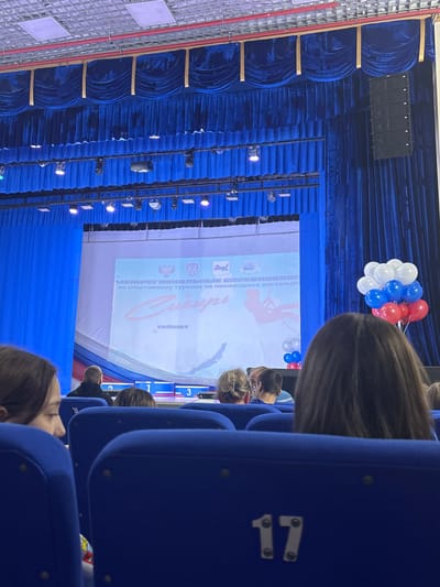 The image depicts the interior of an auditorium in Tayshet, Russia, from an audience perspective, facing a stage. Numerous individuals are seated in blue upholstered chairs, with the back of chair number "17" visible in the immediate foreground. On the stage, a large white projection screen is centrally positioned, displaying text and graphics. The screen shows "МЕЖРЕГИОНАЛЬНЫЕ СОРЕВНОВАНИЯ по спортивному туризму на пешеходных дистанциях" (Interregional Competitions in Sports Tourism on Pedestrian Distances), a stylized "Сибирь" (Siberia), and "ТАЙШЕТ" (Tayshet) at the bottom. The graphics include two silhouettes of individuals rappelling and several logos at the top. Below the screen, a three-tiered winners' podium is visible, marked "1," "2," and "3," decorated with horizontal stripes resembling the Russian flag. Dark blue velvet curtains, some with gold trim, frame the stage. Clusters of red, white, and blue balloons are positioned on both the left and right sides of the stage, near the screen. Above the stage, multiple stage lights, a ceiling-mounted projector, and a vertical array of black speakers are visible. The ceiling consists of a grid system with white panels and exposed red piping. The seated audience members are observing the stage, their backs primarily visible.