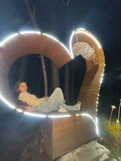 A nighttime, medium-shot photo depicts a young woman in jeans and a beige jacket, positioned within a large, illuminated, Cyrillic letter "Я". The structure appears to be made of wood, edged with bright white lights. The location is Sungurlare, Bulgaria, indicated by Cyrillic script incorporated into the letter's design. The woman is looking at the camera. The surrounding environment is dark.
