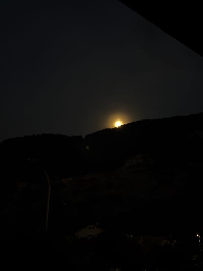 The image depicts a dark nighttime scene in Cluses, France, with a moon or sun partially visible behind a silhouetted mountain range. In the foreground, a dimly lit building with visible windows is situated, along with the outline of a large structure, possibly a crane or wind turbine. The overall ambiance is one of low light and subdued activity, characteristic of a nocturnal environment.