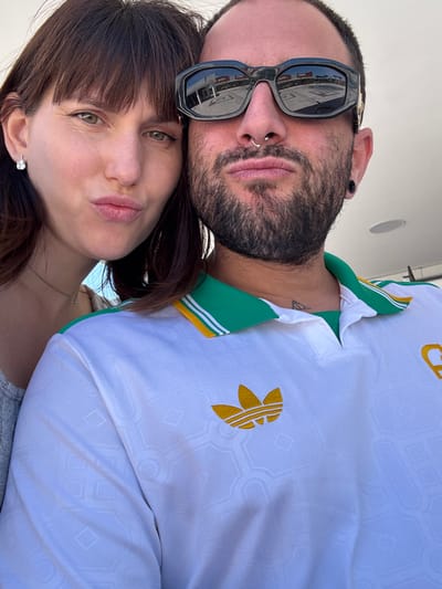 A selfie, taken at an upward angle, shows a man and a woman close together. The man, positioned above, has dark hair, a beard, and sunglasses reflecting a bright urban setting. He also has a nose ring and what appears to be a tattoo on his neck. The woman is below him, with dark hair and a necklace. The man is wearing a white Adidas shirt with green and yellow accents. The background suggests an outdoor location, possibly in Pomezia, Italy, based on reflections in the man's sunglasses.