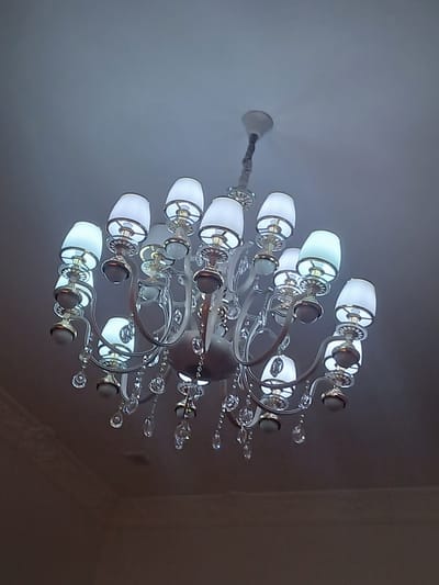 A chandelier with multiple arms hangs from a ceiling. The chandelier is decorated with crystal accents and has white lampshades. The background is a plain, light-colored ceiling. The image appears to be taken indoors.