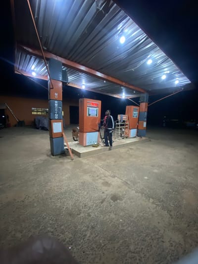 The image depicts a nighttime scene at a gas station in Sasaro, Nigeria. A male individual, clad in a dark jacket with white arm stripes and dark trousers, stands on a raised concrete platform beside an orange and white fuel pump. He is actively holding a fuel nozzle, appearing to dispense fuel. This pump displays red digital numbers and a screen below, where text including '8229950428' and 'BOSS PHONE' is discernible. A second, identical fuel pump is positioned to the left, partially obscured, with a 'Lubricants' label visible.
An orange-brown metal framework supports a corrugated metal canopy overhead, from which multiple bright white lights illuminate the station. Centrally located, a tall pillar is constructed from alternating orange-brown and dark gray rectangular blocks. The dark gray sections feature vertical white text stating 'NO SMOKING'. A small sign is affixed to an orange block, and a slender orange-brown rod leans against the pillar's base.
In the background to the left, a building with an orange-brown facade and gridded windows is visible, with a long pole or ladder leaning against it and tarp-covered items nearby. The ground is a textured concrete surface with visible cracks and variations in color. The areas beyond the station's direct illumination are dark.
