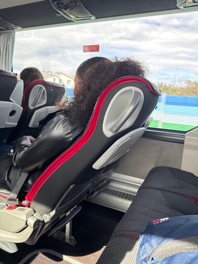 The scene depicts the interior of a passenger coach or bus, observed from a forward-facing perspective. A person with dark, curly hair, pulled up and away from the neck, is seated with their back to the viewer. They are wearing a black, shiny jacket, possibly leather or faux leather, and a seatbelt is visible across their lap. Their seat is predominantly black fabric with red piping accents, featuring a grey plastic handle and a folded-up tray table mechanism on its back. To the left and slightly forward, another passenger with brown hair is partially visible in an adjacent seat.
Through the large window on the right side of the vehicle, a view of the exterior shows a partly cloudy sky. In the middle distance, residential buildings with light-colored roofs are visible against the horizon. Closer to the vehicle's path, a long structure composed of horizontal blue and green panels is present, suggesting a barrier or building alongside the road. A red rectangular sticker with a white 'X' mark is affixed to the window glass. The floor of the vehicle is dark. In the lower right foreground, a blue backpack with a "TOMMY" brand label is resting on an unoccupied seat. The vehicle is currently situated in or passing through the city of Macedo de Cavaleiros, Portugal.