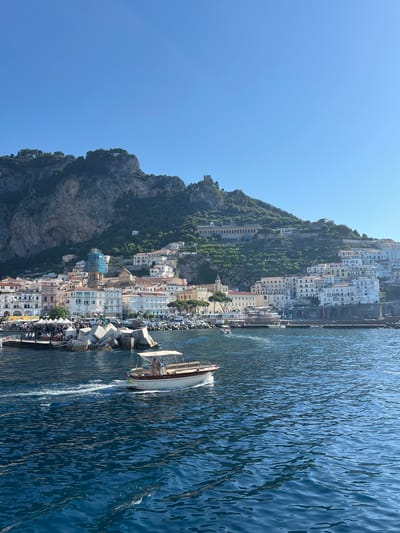 An eye-level, long shot captures a bright, sunny day with a clear blue sky over the coastal city of Pogerola, Italy. The city, built on a steep hillside, is densely packed with white and cream-colored buildings. Terraced gardens and trees are visible on the slopes behind the city. A small boat with a canopy and two people on board is in the foreground, moving from left to right across the deep blue water. Other boats are docked in the harbor. A breakwater made of large concrete blocks is also visible. The scene appears peaceful and picturesque, with the natural beauty of the landscape complementing the architecture of the city.