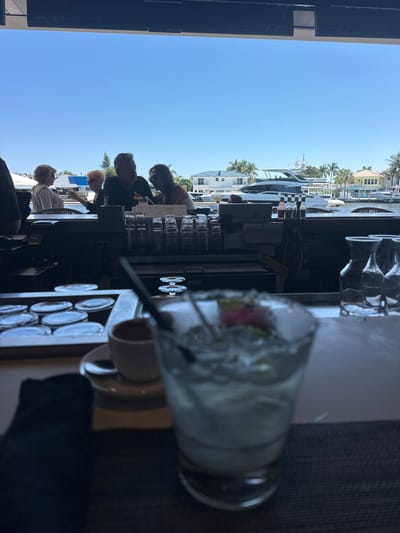 The image depicts a bright daytime scene from inside a restaurant or bar, looking out onto a waterway in Lauderdale-by-the-Sea, United States of America.
In the immediate foreground, a blurred cocktail glass with a sugared rim, ice, a dark straw, and a red and green garnish (possibly fruit) is prominent on a dark tabletop. To its left, a small white ceramic cup and saucer with a spoon are visible, along with a dark folded napkin. Behind these items, on what appears to be a bar counter, there are stacked clear glasses, a tray containing multiple round, lidded containers, various small bottles, and several clear pitchers.
In the midground, silhouetted against the bright outdoor light, four individuals are seated at a bar or table. On the left, two older women appear to be conversing. To their right, a man in a dark shirt and a woman in a light top are engaged in conversation; the man holds a drink.
The background reveals a wide, calm waterway under a clear, bright blue sky. Several boats are docked along the water's edge, including a large white yacht with a dark checkered pattern on its hull, and another white yacht further in the distance. Across the waterway, residential houses with light-colored walls and dark roofs are visible, interspersed with numerous green palm trees, consistent with a coastal subtropical environment. The overall lighting suggests direct sunlight outdoors.