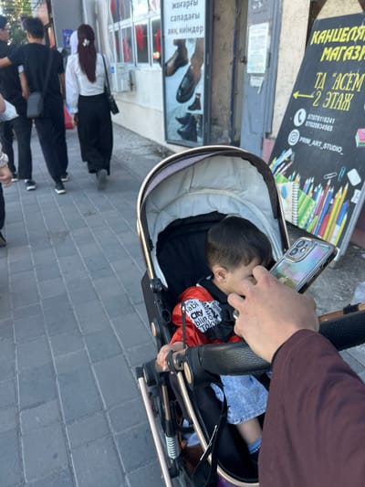 A photo shows a baby lying in a stroller. A person takes a photo with a phone. People walk on the sidewalk. A store sign advertises stationery, stating it is located in Aksu, Kazakhstan. The sign also lists phone numbers and the Instagram handle for "PRIM_ART_STUDIO." Another sign promotes high-quality footwear. The ground is paved with gray bricks.