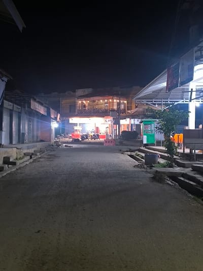 The image depicts a nighttime street scene in the city of Manetunong, Indonesia. The perspective is from the center of a dark, asphalt road looking forward towards a series of buildings. On the left, a row of single-story structures with closed metal shutters appears mostly dark, with a single bright light source illuminating a small section. Directly ahead and slightly to the right, a two-story building features a brightly lit upper balcony area with seating and signage, including the word "KOPI." Below the balcony, the ground floor is also brightly illuminated. Approximately eight motorcycles are parked in front of this building, partially obscured by red barriers. To the far right, a large white awning structure covers an open area, brightly lit by white fluorescent lights. Under this awning, a small green kiosk stands beside a young tree, and several orange plastic chairs with a table are visible. The sky above is completely dark, indicating night. No individuals are clearly visible in the scene. The road surface is uneven, with visible cracks and worn pavement on the edges, and concrete steps leading up to the buildings.